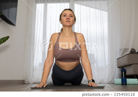 Woman Performs Cobra Pose on Yoga Mat in Bright Living Room Woman Performs Cobra Pose on Yoga Mat in Bright Living Room 135450118