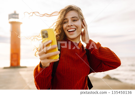 Young woman smiling and using her smartphone at the beach sunset. Blogging, communication concept. Young woman smiling and using her smartphone at the beach sunset. Blogging, communication concept. 135450136
