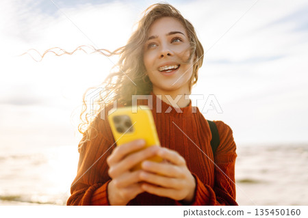 Young woman using mobile phone at beach. Cold weather. Autumn. Blogging, communication Young woman using mobile phone at beach. Cold weather. Autumn. Blogging, communication 135450160