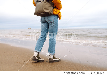 Woman traveler walks in boots along the seashore. Happy traveler with a backpack enjoys the sea air. Woman traveler walks in boots along the seashore. Happy traveler with a backpack enjoys the sea air. 135450424