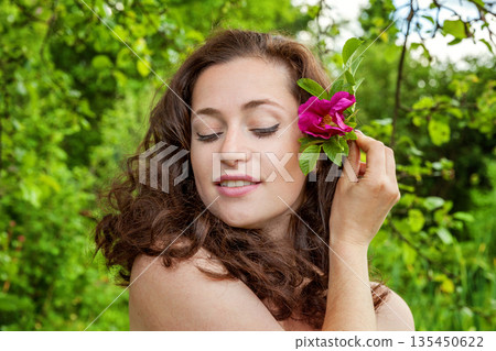 Beautiful young brunette girl with perfect skin holding red rose flower in hand on park or garden green background. Beauty nature skincare eco organic cosmetic ecology pure health concept 135450622