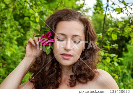 Beautiful young brunette girl with perfect skin holding red rose flower in hand on park or garden green background. Beauty nature skincare eco organic cosmetic ecology pure health concept 135450623