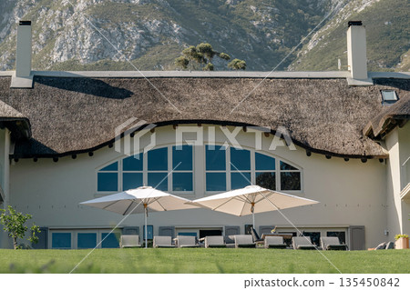 Luxury home with thatched roof and patio chairs on a lawn with mountains 135450842