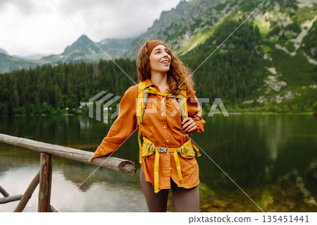 Travel Slovakia, Europe. Tourist with a yellow backpack stands against the backdrop of alpine lake. Travel Slovakia, Europe. Tourist with a yellow backpack stands against the backdrop of alpine lake. 135451441