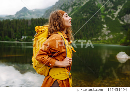 Travel Slovakia, Europe. Tourist with a yellow backpack stands against the backdrop of alpine lake. Travel Slovakia, Europe. Tourist with a yellow backpack stands against the backdrop of alpine lake. 135451494