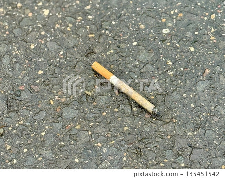 Cigarette Butt On Rough Asphalt, CloseUp, Discarded Filter With Smoldering Ash, Granular Pavement Texture With Small Stones, Faded Paint Specks, Urban Parking Lot Atmosphere, Littering 135451542