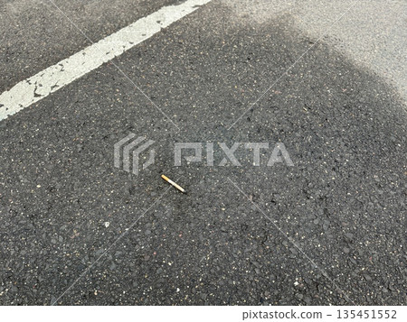 Cigarette Butt On Rough Asphalt, CloseUp, Discarded Filter With Smoldering Ash, Granular Pavement Texture With Small Stones, Faded Paint Specks, Urban Parking Lot Atmosphere, Littering 135451552