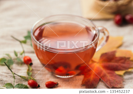 Glass cup of red herbal tea with rosehip berries on autumn leaves, cozy seasonal still life with warm fall atmosphere. 135452237