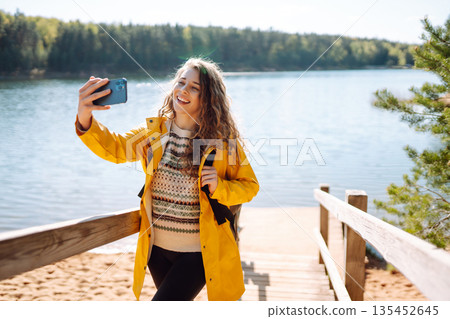 Happy woman with phone in hands taking selfie in forest. Young tourist enjoying spring day near lake 135452645