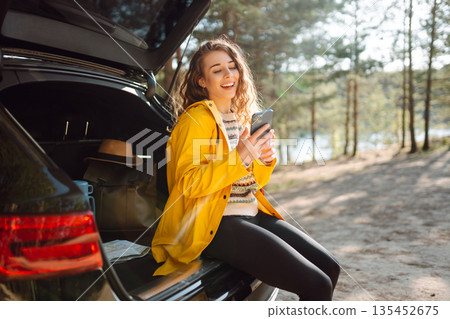 Young woman sitting in open trunk with phone. Lifestyle, travel, tourism, nature, active life. 135452675