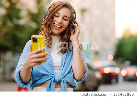 Happy relaxed woman in wireless headphones choosing, listening favorite music in urban city street. 135452890