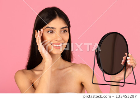 Young smiling woman touching face while holding mirror in studio 135453297
