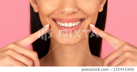 Closeup smile of young beautiful woman pointing at white teeth on pink background Closeup smile of young beautiful woman pointing at white teeth on pink background 135453308