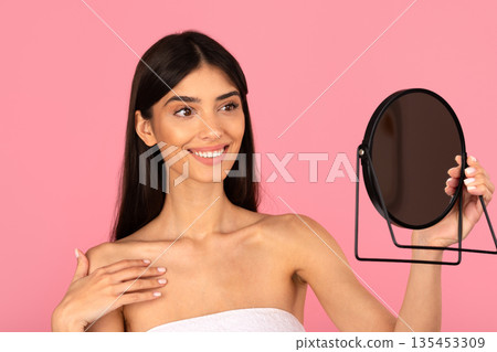 Young beautiful woman smiling and looking in handheld mirror on pink background Young beautiful woman smiling and looking in handheld mirror on pink background 135453309