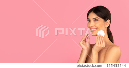 Smiling young woman using cotton pads on face during daily skincare routine 135453344