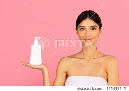 Young woman presenting cosmetic bottle on open palm against pink background 135453345