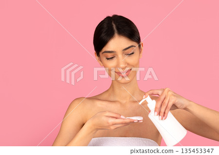 Young adult woman dispensing skincare lotion onto cotton pad from pump bottle Young adult woman dispensing skincare lotion onto cotton pad from pump bottle 135453347