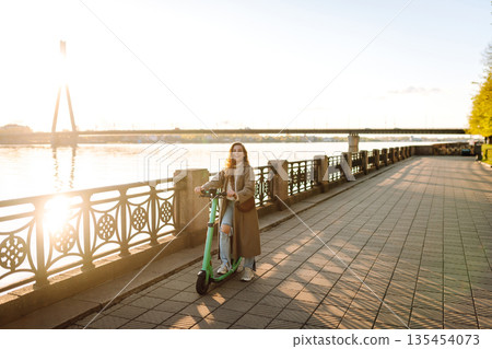 Young woman enjoys riding electric scooter along waterfront promenade with view. Travel concept. 135454073