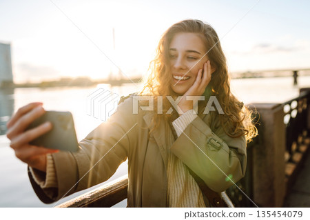 Selfie time. Woman holding mobile phone taking selfie photo, standing on waterfront. Lifestyle. 135454079