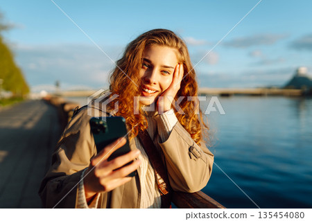 Selfie time. Woman holding mobile phone taking selfie photo, standing on waterfront. Lifestyle. 135454080