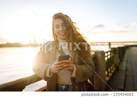 Young woman enjoys a sunny day by the river, using her smartphone while smiling in the warm sunlight 135454081