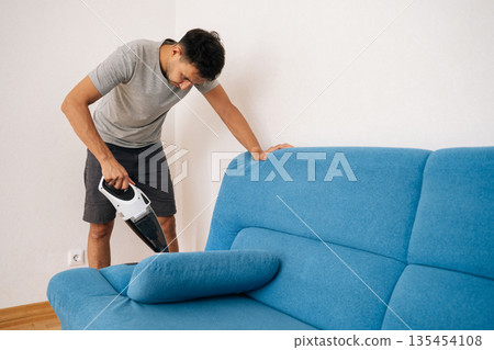 Portrait of focused young man using handheld vacuum cleaner to remove dust and dirt from blue sofa, emphasizing importance of cleanliness and hygiene in home maintenance. 135454108