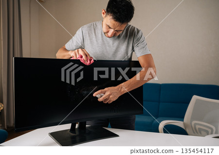 Careful cleaning of curved computer monitor screen using disinfectant spray and microfiber cloth, maintaining hygienic workspace for optimal working conditions. Concept of household cleanliness. Careful cleaning of curved computer monitor screen using disinfectant spray and microfiber cloth, maintaining hygienic workspace for optimal working conditions. Concept of household cleanliness. 135454117