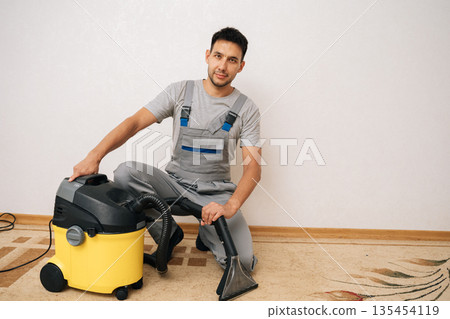 Portrait of handsome skilled cleaner male sitting on floor next to washing vacuum cleaner, holding cleaning attachment, prepared to deliver expert carpet cleaning services in home environment 135454119