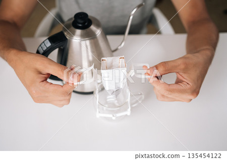 Top view of unrecognizable coffeemaker placing drip coffee bag onto glass cup, showcasing process of brewing freshly ground coffee with kettle in background. Concept of enjoying morning ritual. Top view of unrecognizable coffeemaker placing drip coffee bag onto glass cup, showcasing process of brewing freshly ground coffee with kettle in background. Concept of enjoying morning ritual. 135454122
