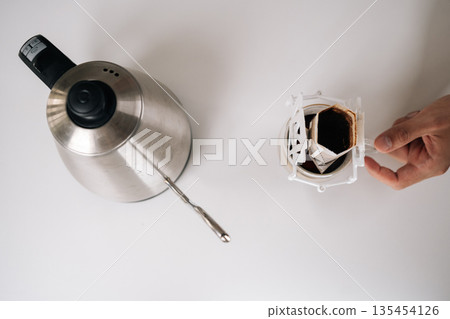 Coffee brewing process, male hand pouring hot water from gooseneck kettle onto paper filter, glass dripper placed on minimalist white surface, soft kitchen lighting. Concept of enjoying morning ritual 135454126