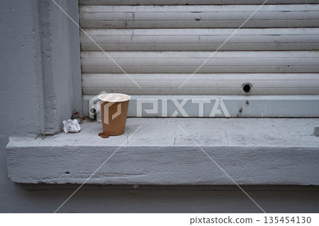 Cardboard coffee cup on a window sill next to coins and a crumpled receipt. Casual everyday scene showing disposable items and small personal objects in an outdoor setting. 135454130
