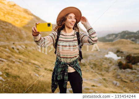 Young woman with backpack is happily taking selfie in mountains. Freedom adventure, happy vacation. Young woman with backpack is happily taking selfie in mountains. Freedom adventure, happy vacation. 135454244