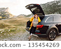 A cheerful traveler enjoying a scenic mountain view while sitting on the trunk. Travelling by car. 135454269