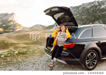 A cheerful traveler enjoying a scenic mountain view while sitting on the trunk. Travelling by car. A cheerful traveler enjoying a scenic mountain view while sitting on the trunk. Travelling by car. 135454269