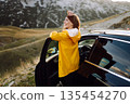 A cheerful traveler enjoying a scenic mountain view while sitting on the trunk. Travelling by car. 135454270