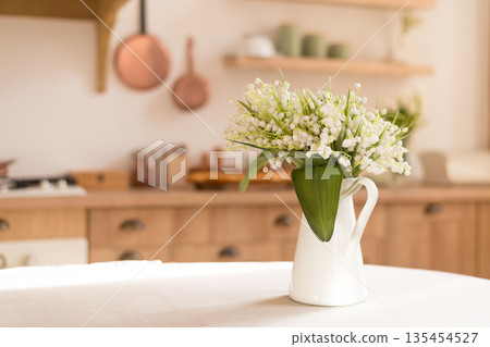 Fresh bouquet of lilies of the valley in a white ceramic jug on a table. Cozy kitchen interior with wooden furniture in the blurred background. Fresh bouquet of lilies of the valley in a white ceramic jug on a table. Cozy kitchen interior with wooden furniture in the blurred background. 135454527