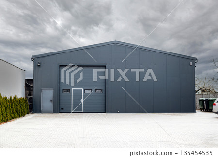 Modern gray metal warehouse building with a large sectional garage door under a cloudy sky. Industrial storage facility. 135454535