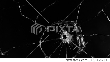 Front view photo of glass shattered by a bullet. Gunshot impact creates cracks, flying shards and dust isolated on black background. Perfect VFX overlay element or dramatic background. 135454711