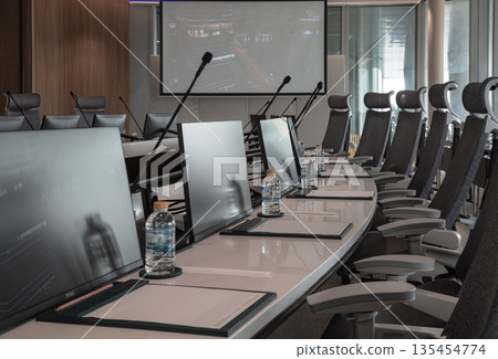 A sleek, modern boardroom features a curved white table lined with ergonomic grey chairs. Each participant's station is individually equipped with a slim monitor, a gooseneck microphone, and a bottle A sleek, modern boardroom features a curved white table lined with ergonomic grey chairs. Each participant's station is individually equipped with a slim monitor, a gooseneck microphone, and a bottle 135454774