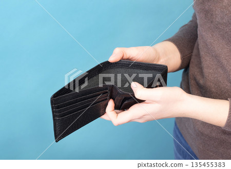 Person holding and opening an empty black leather wallet against a blue background. Person holding and opening an empty black leather wallet against a blue background. 135455383