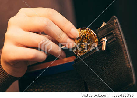 Hand putting a golden bitcoin coin into a black leather wallet, representing crypto savings and digital currency. 135455386
