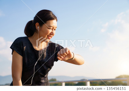 Active Asian woman checking her fitness tracker after a run outdoors. Concept of workout recovery, performance monitoring, healthy living, self care and active lifestyle. 135456151