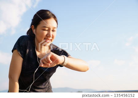 Active Asian woman checking her fitness tracker after a run outdoors. Concept of workout recovery, performance monitoring, healthy living, self care and active lifestyle. 135456152