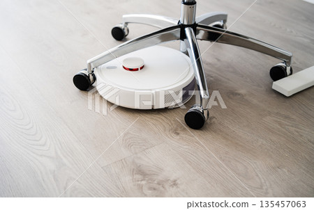Robot vacuum cleaner cleaning under office chair on wooden floor in modern home interior, smart automated household technology Robot vacuum cleaner cleaning under office chair on wooden floor in modern home interior, smart automated household technology 135457063