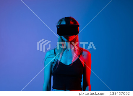 Young woman wearing virtual reality headset standing in studio with blue and red lighting, immersive technology concept Young woman wearing virtual reality headset standing in studio with blue and red lighting, immersive technology concept 135457092