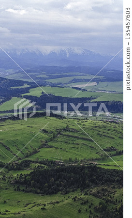 Vast green meadows stretch into the distance with snow-capped mountains rising under a cloudy sky. Vast green meadows stretch into the distance with snow-capped mountains rising under a cloudy sky. 135457603