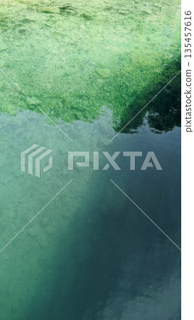 Top-down view of transparent turquoise water revealing stones beneath the surface, with trees reflected in the calm upper half. Top-down view of transparent turquoise water revealing stones beneath the surface, with trees reflected in the calm upper half. 135457616