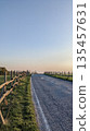 Empty rural road in Italy at golden hour, bordered by wooden fences and grassy fields, leading toward the horizon under a clear evening sky. 135457631
