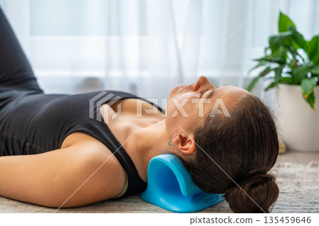 Young woman lies on the floor using a blue cervical traction device to stretch the neck. Home therapy for neck pain, muscle tension and spine decompression. Young woman lies on the floor using a blue cervical traction device to stretch the neck. Home therapy for neck pain, muscle tension and spine decompression. 135459646