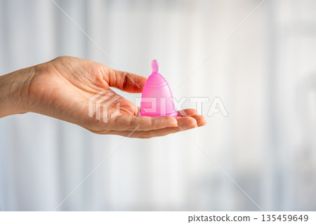 Close-up of a pink menstrual cup standing upright on a woman open palm over wooden table. Feminine hygiene product promoting zero waste, comfort, and sustainable period care. 135459649
